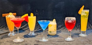 Renkli mocktail ve soğuk içeceklerden oluşan canlı bir seçim. Zarif cam örtülerde koyu dokulu arka planda servis ediliyor..