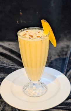 Dilimlenmiş bademlerle kaplı ve taze mangolarla süslenmiş uzun bir bardakta servis edilen kremalı mangolu smoothie. Yaz içeceği menüleri, sağlıklı içecek promosyonları ve gıda pazarlaması için mükemmel..