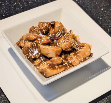 Zengin kahverengi bir sosla kaplanmış susamlı tavuğun yakın çekimi ve üstü kızarmış susam tohumlarıyla kaplı, modern kare beyaz bir kasede servis ediliyor..