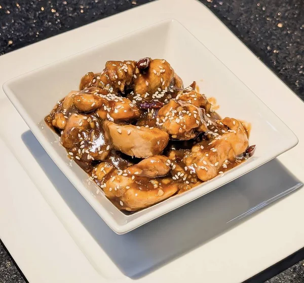 Zengin kahverengi bir sosla kaplanmış susamlı tavuğun yakın çekimi ve üstü kızarmış susam tohumlarıyla kaplı, modern kare beyaz bir kasede servis ediliyor..