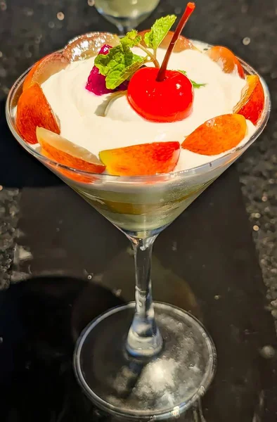 Üstü krema, kiraz, nane ve taze şeftali dilimleriyle kaplı martini bardağında servis edilen zarif katmanlı meyve tatlısı..