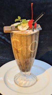 Görsel olarak zengin ve çikolata aromalı bir milkshake veya smoothie, uzun, saplı bir bardakta sunuluyor, iç muz dilimleri ve kalın bir dokuya sahip.