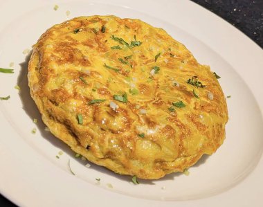 Taze pişmiş patates omleti, altın sarısı, kabarık bir dokuya sahip, otlarla süslenmiş ve temiz beyaz bir tabakta servis edilmiş..