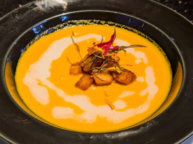 Zengin, kadife kabak çorbası koyu bir kasede servis ediliyor, kremayla güzelce kıvrılmış ve üzerinde altın kıtır ekmekler ve kızarmış otlar var..