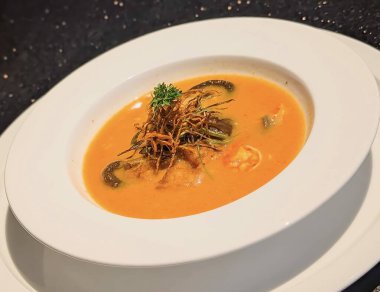 Karides, mantar ve canlı portakal suyuna batırılmış taze sebzelerin yer aldığı koyu beyaz bir kasede sunulan zengin ve aromatik bir deniz ürünleri çorbası..