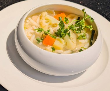 Kalın beyaz erişteler, taze karnabahar, havuç ve yeşil soğan içeren parlak, berrak bir erişte çorbası, eşleşen beyaz bir tabağın üzerinde pipo sıcaklığında servis edilir..