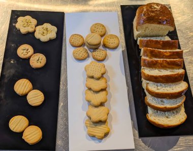 Çeşit çeşit tereyağlı kurabiyeler ve zıt siyah ve beyaz tabaklar üzerinde sunulan taze dilimlenmiş beyaz ekmek, dokusunu, rengini ve fırın tarzı basitliğini gözler önüne seriyor..