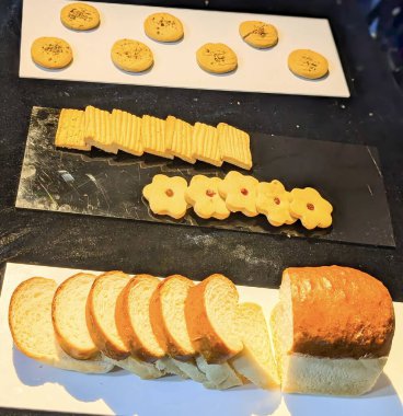 Düzenlenmiş çeşitli kurabiyeler ve taze dilimlenmiş yumuşak ekmekler sıcak ışık altında beyaz ve siyah servis malzemeleriyle sunuluyor..