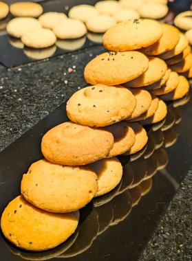 : Dikey, yakın plan çekimde birden fazla taze pişmiş, altın renkli kurabiye yığını ele geçirilir, özellikle üzerinde siyah susam tohumları olanlar, son derece yansıtıcı siyah bir tabakta çapraz olarak sıralanır..