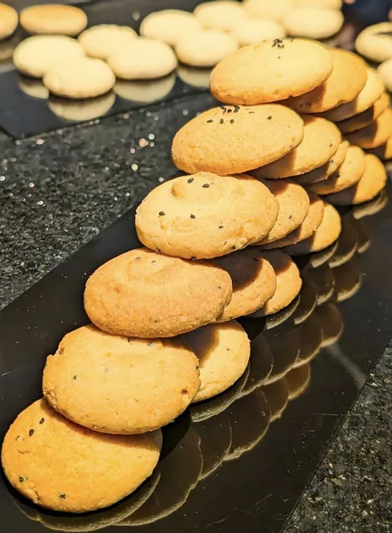 : Dikey, yakın plan çekimde birden fazla taze pişmiş, altın renkli kurabiye yığını ele geçirilir, özellikle üzerinde siyah susam tohumları olanlar, son derece yansıtıcı siyah bir tabakta çapraz olarak sıralanır..