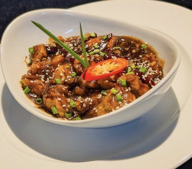 Lezzetli Mançurya tavuk parçalarının yakın plan çekimi zengin, koyu jöleli, kızarmış susam tohumu, doğranmış soğan ve bir dilim kırmızı biber ile süslenmiş..