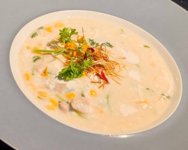 Taze otlarla süslenmiş zengin ve kremalı tavuk çorbası, çıtır çıtır iplikler ve yenilebilir çiçek yaprakları. Gri çerçeveli tabakla beyaz bir kasede sunuluyor..