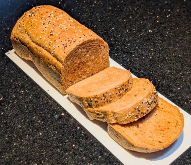 Üzerinde susam ve karışık tohumlar bulunan bir somun çoklu tahıllı ekmek, kısmen dilimlenmiş ve koyu bir tezgahın üzerinde beyaz bir dikdörtgen tepsiye yayılmış..