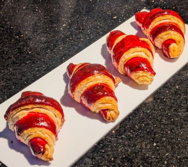 Yakın plan altın-kahverengi kruvasan, canlı kırmızı jöle ya da çizgili dolgu, beyaz servis tepsisi üzerine dizilmiş.