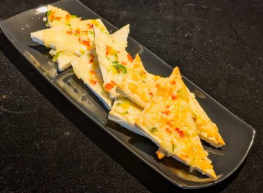 Eritilmiş peynir ve ince doğranmış dolmalık biberle kaplı, sade siyah bir tabakta servis edilen altın tost ekmeği üçgenleri.