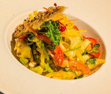 Lezzetli soslu salatalık, domates, dolma biber ve kök sebzelerden oluşan canlı bir karışım, beyaz bir tabakta kavisli beyaz bir kasede servis ediliyor..