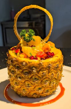 İçinde portakal dilimleri, papaya parçaları, nar tohumları, mısır çekirdekleri ve taze bitkiler bulunan içi oyulmuş ananas kabuğunda servis edilen renkli bir tropikal meyve salatası..