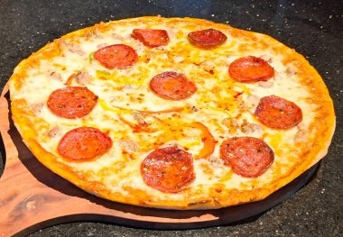 Yeni pişmiş bir pizza, erimiş peynir, pepperoni dilimleri, sosis parçaları ve kırmızı ve yeşil çan biberleriyle kaplı, ahşap bir tahtanın üzerinde servis edilir..