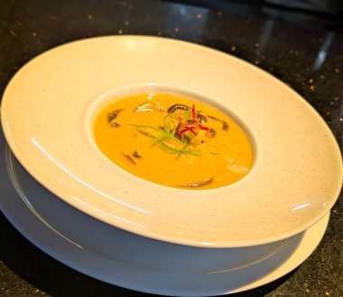 Dilimlenmiş mantar, yeşil soğan ve kırmızı biber şeritleri içeren Tayland esinlenilmiş bir portakal çorbası kasesi, beyaz seramik ile koyu bir arka fonda servis edilir..