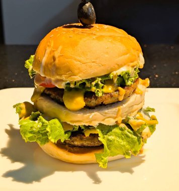 İki etli çizburger, erimiş çedar, ızgara soğan ve kavrulmuş biber, susamlı çöreğin üzerinde servis edilmiş ve kırmızı biber ve zeytinle süslenmiş..