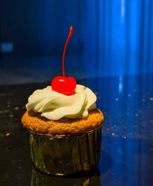 Metalik bir ambalajda altın bir kek pürüzsüz bir krema ve parlak kırmızı bir kiraz ile kaplanır. Derin mavi bir arka planda dramatik ışık altında yakalanır..