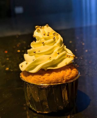 Pürüzsüz, altın renkli krema ve metal şeker incileriyle kaplı zengin bir vanilyalı kek zarif, kaliteli bir görünüm için yansıtıcı folyo bardakta sunuluyor..