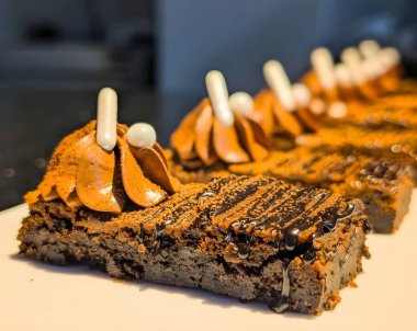 Beyaz dikdörtgen bir tabakta servis edilen krema, beyaz çikolata çubukları ve şeker incileri ile kaplı zarif bir sıra çikolatalı tatlı..