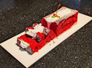 Çırpılmış kremayla kaplanmış ve kirazla süslenmiş canlı kırmızı kadife kek, nemli dokusunu ve zengin kırıntılarını ön plana çıkaran temiz beyaz bir tabakta sunuluyor..