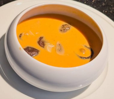 Dilimlenmiş mantarlar ve zengin kremalı dokularla modern beyaz bir kasede servis edilen canlı portakal çorbası..