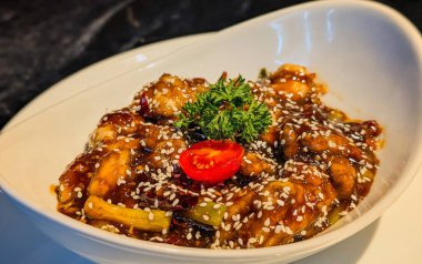 Isırık büyüklüğünde, parlak koyu kahverengi sosla kaplanmış, susam tohumları, yeşil soğan, kurutulmuş kırmızı biber, maydanoz ve vişneli domatesle süslenmiş tavuk parçaları..