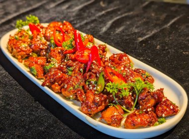 Isırık büyüklüğünde, kırmızımsı kahverengi sosla kaplanmış, susam tohumları, kırmızı biber dilimleri ve taze otlarla süslenmiş tavuk parçaları..