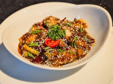 Isırık büyüklüğünde, parlak koyu kahverengi sosla kaplanmış, susam tohumları, yeşil soğan, kurutulmuş kırmızı biber, maydanoz ve vişneli domatesle süslenmiş tavuk parçaları..