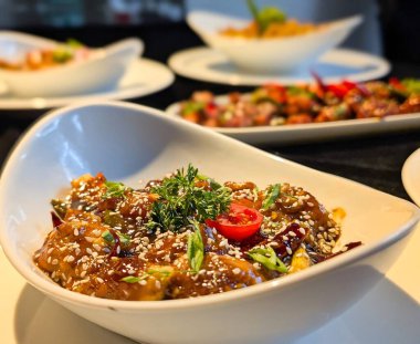 Isırık büyüklüğünde, parlak koyu kahverengi sosla kaplanmış, susam tohumları, yeşil soğan, kurutulmuş kırmızı biber, maydanoz ve vişneli domatesle süslenmiş tavuk parçaları..