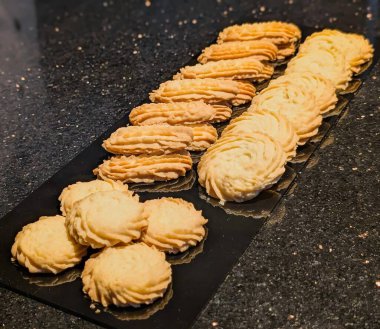 Sıralı tereyağlı mermiler, susamlı tepsiler, borulu altın kurabiyeler ve pürüzsüz garnitürlü çeşitleri içeren çeşitli kurabiye çeşitleri ayrı tepsilerde dizilmiştir.