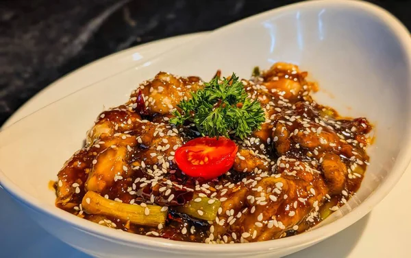 Isırık büyüklüğünde, parlak koyu kahverengi sosla kaplanmış, susam tohumları, yeşil soğan, kurutulmuş kırmızı biber, maydanoz ve vişneli domatesle süslenmiş tavuk parçaları..