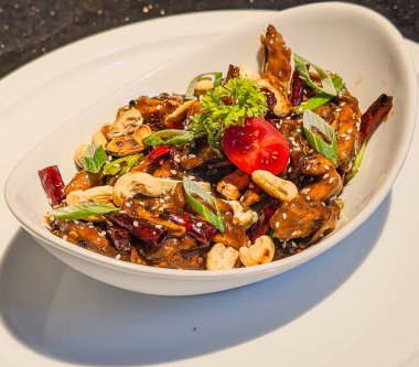 Klasik Kung Pao tavuğu, içinde susam tohumları, yeşil soğan, kurutulmuş kırmızı biber, kaju fıstığı ve vişne domatesli jöleli tavuk parçaları bulunan beyaz oval bir kasede servis edilir.