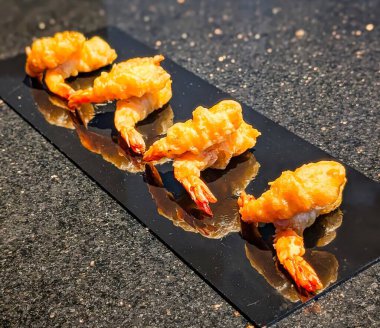 Pürüzsüz, siyah dikdörtgen bir tabakta sıralanmış gevrek altın karides tempurası. Açık kuyruklar ve havadar vurucular sergiliyor..