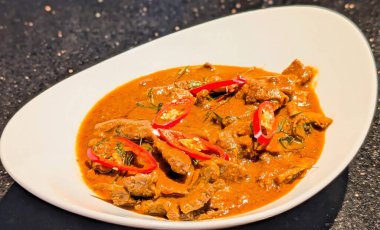 Beyaz oval tabakta servis edilen ve kırmızı biber ve kaffir limon yapraklarıyla süslenmiş yumuşak et dilimli zengin Tayland kırmızı körisi..