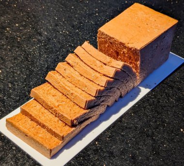 Kısmen dilimlenmiş turuncu ekmek beyaz dikdörtgen bir tabakta koyu bir tezgahın üzerinde sergileniyor..