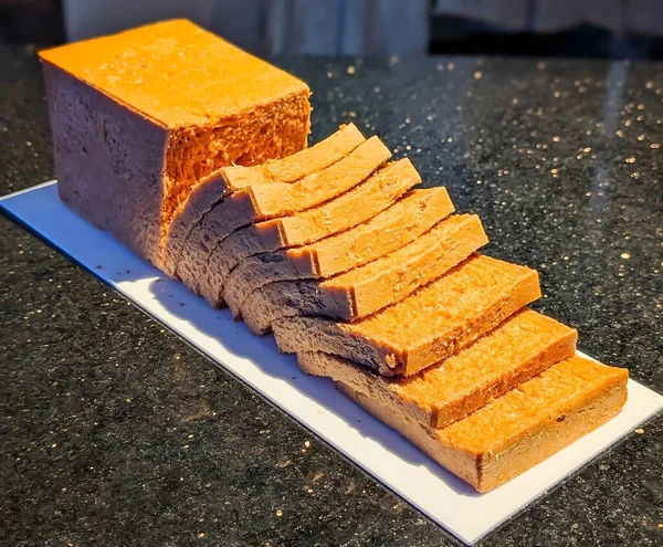 Kısmen dilimlenmiş turuncu ekmek beyaz dikdörtgen bir tabakta koyu bir tezgahın üzerinde sergileniyor..