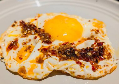 Beyaz bir tabakta mükemmel bir şekilde kızartılmış yumurta, altın sarısı ve çıtır çıtır kenarlı, üzerinde canlı kırmızı biber yağı ve kırmızı biber taneleri var..