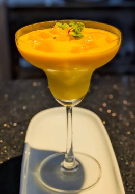 Margarita tarzı bardakta servis edilen kalın mango püresi. Üstünde mango parçaları ve taze nane filizi var..
