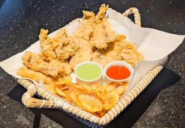 Kağıt havlularla dolu bir sepette servis edilen altın sarısı fileto ve patates cipsi, yanında da yeşil ve kırmızı sos..