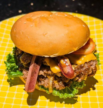 Çift köfteli gurme, erimiş çedar peyniri, marul, domates, ızgara soğan ve dilimlenmiş sosis içeren bir hamburgerin yakın çekimi sarı ızgara desenli tabakta servis edilir..