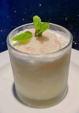 Köpüklü hindistan cevizi sütü, köpüklü tatlı, pembe granüller ve taze bir nane filizi, beyaz bir tabakta servis edilen temiz bir bardakta..