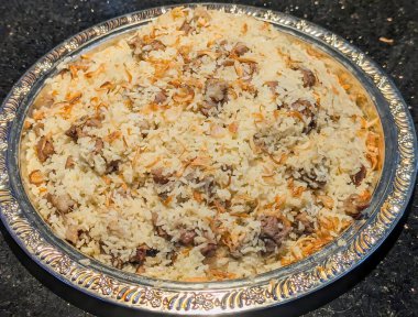 Süslü kuzu biryani tabağı kabarık safran pirinç ve karamelli kızarmış soğan, gümüş servis tepsisine dizilmiş sıcak yönlü aydınlatma ve parlak koyu arkaplan.