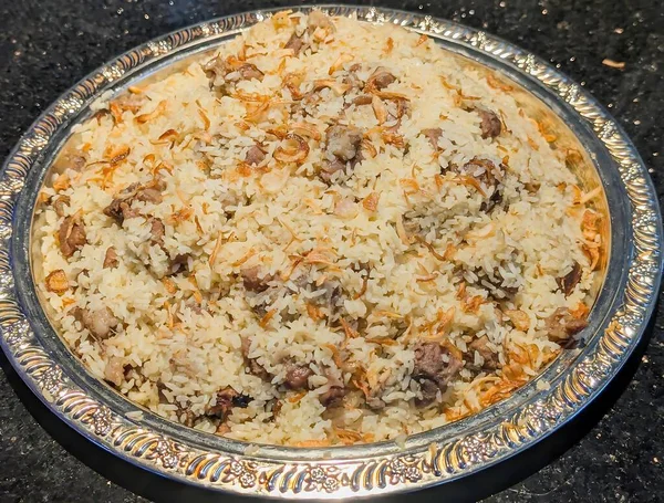 Süslü kuzu biryani tabağı kabarık safran pirinç ve karamelli kızarmış soğan, gümüş servis tepsisine dizilmiş sıcak yönlü aydınlatma ve parlak koyu arkaplan.