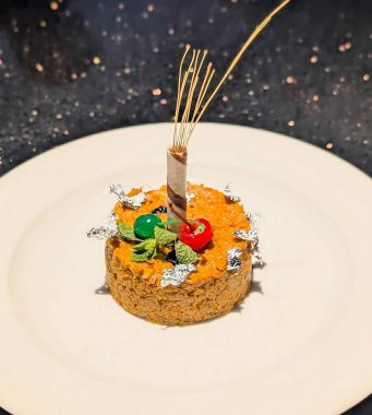 Silindirik fındıklı halwa üzerine yenebilir gümüş folyo, dikey çikolata gofreti, jöleli kiraz ve taze nane şekeri, beyaz bir tabakta sunulur..