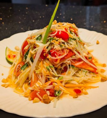 Bu canlı ve otantik Tayland yeşil papaya salatası (Som Tum) parçalanmış papaya, taze kiraz domatesleri ve dekoratif beyaz bir tabakta çıtır fıstık içerir.