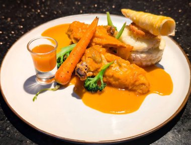 Parlak portakal soslu gurme tavuk, katmanlı patates yığını, kızarmış havuç, brokoli çiçekleri ve yanında beyaz bir tabakta bir bardak sos eşliğinde rulo pastayla kaplıdır..